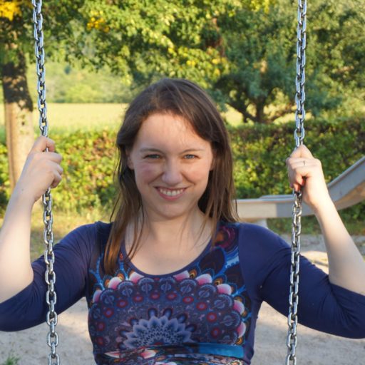 Isabella Benz sitzt auf einer Schaukel und sieht geradewegs in die Kamera. Ihre Haare sind offen. Sie hält sich mit beiden Händen auf Höhe ihres Kopfes an der Schaukel fest. Sie trägt ein dunkelblaues Kleid mit einem Mandala-Muster. Die Ärmel reichen bis zu den Ellenbogen. Im Hintergrund sieht man eine Rutsche und eine grüne Hecke.